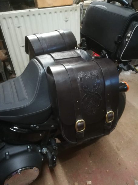Leather saddlebag attached to a motorcycle seat with decorative stitching.
